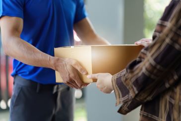 Person in plaid shirt receives boxed package from delivery person in blue shirt.