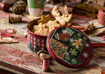 homemade delicious gingerbread cookies in vintage gift round metal box with christmas ornaments on wooden rustic red table with cup of coffee with foam, christmas tree branches opposite brick wall