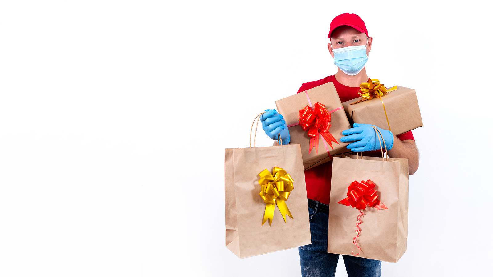 Safe contactless remote delivery of holiday gifts during coronavirus pandemic. A courier in red uniform and protective medical mask and gloves holds large order, many gift boxes and bags with bows